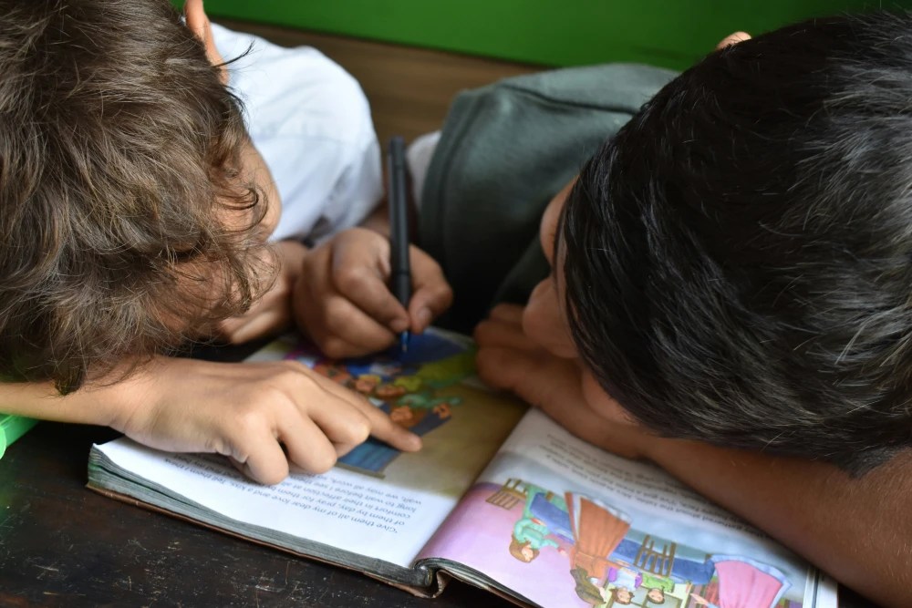two boys, huddled closely together look at a book. they seem to be working together, with one pointing at the page and the other writing.
